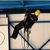 Průmyslový lezec ve visu na laně pod modrou ocelovou konstrukcí haly